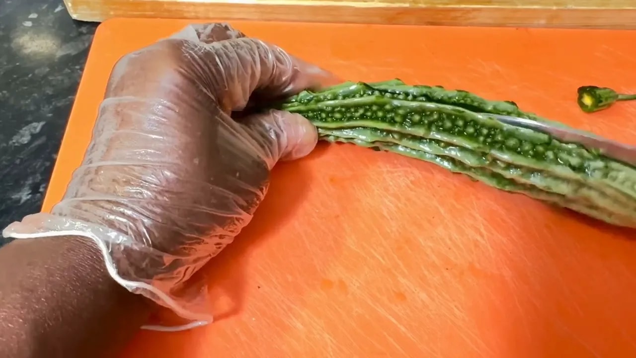 Gloved hand holding bitter melon halves on an orange cutting board during prep